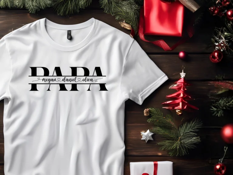 White crewneck t-shirt with a split monogram design featuring 'PAPA' in bold black serif letters with the names 'megan ♡ daniel ♡ oliva' in cursive script running through the center. Shirt is flat-laid on a dark wood surface styled with Christmas decorations including pine branches, red ornaments, wrapped gifts with red ribbon, and a decorative red paper Christmas tree.
